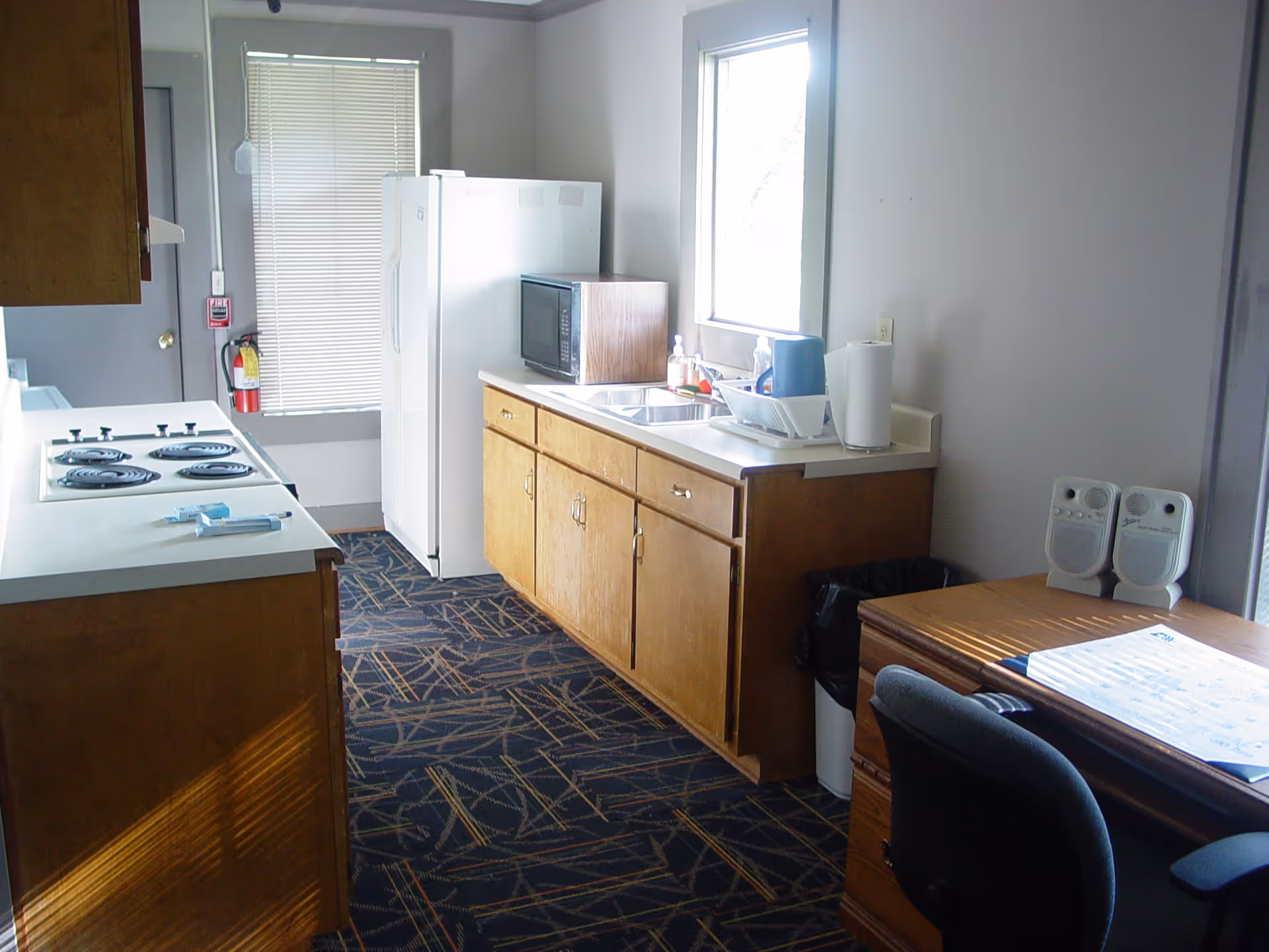 A small kitchen area with wooden cabinets, a white electric stove, a white refrigerator, a microwave on the counter, a sink with dish soap and a drying rack, a window with blinds, a fire extinguisher mounted on the wall, and a desk with a chair and two small speakers.
