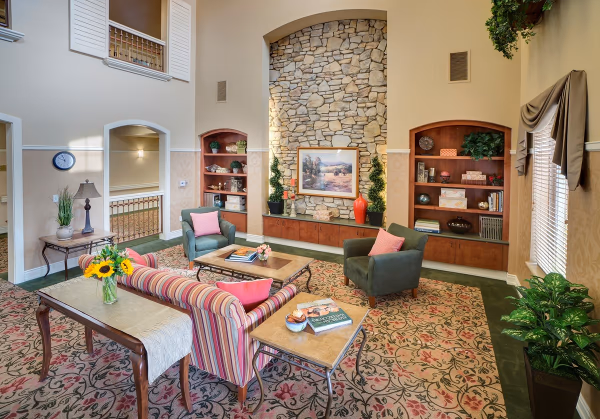 A cozy living room area in Eskaton Village Roseville featuring a stone fireplace with a framed landscape painting above it. The room has two green armchairs with pink pillows, a striped sofa with pink pillows, a wooden coffee table with books and flowers, and a side table with a book and a bowl. Built-in wooden shelves flank the fireplace, decorated with plants, books, and decorative items. The room has a floral patterned carpet, beige walls, and a window with blinds and a valance. There are also plants and a small table with a lamp and a clock on the wall.