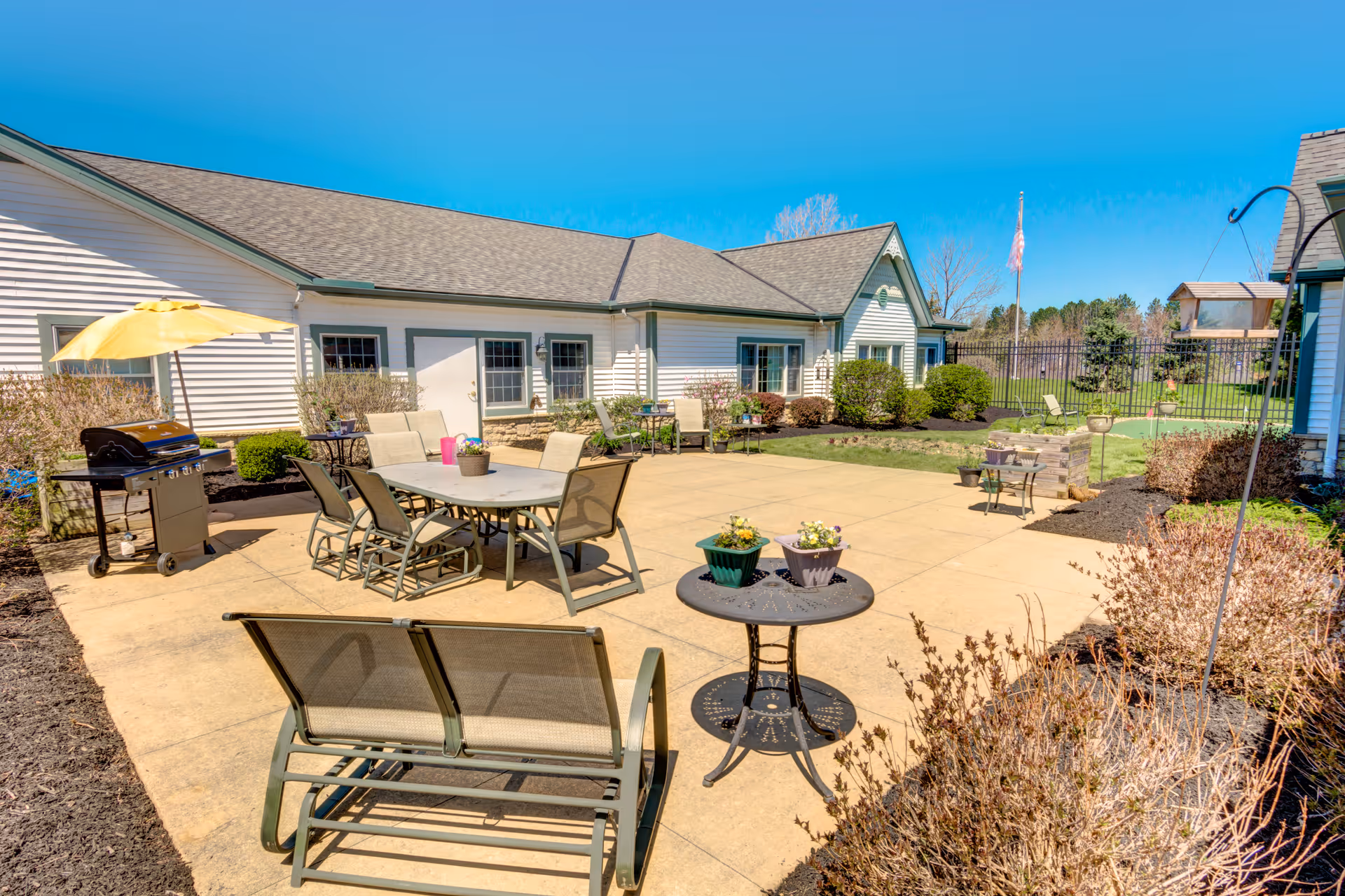 Outdoor patio area at Symphony at Mentor with multiple seating arrangements including tables and chairs, a grill with a yellow umbrella, potted plants on tables, and a well-maintained garden with shrubs and a flagpole in the background under a clear blue sky.