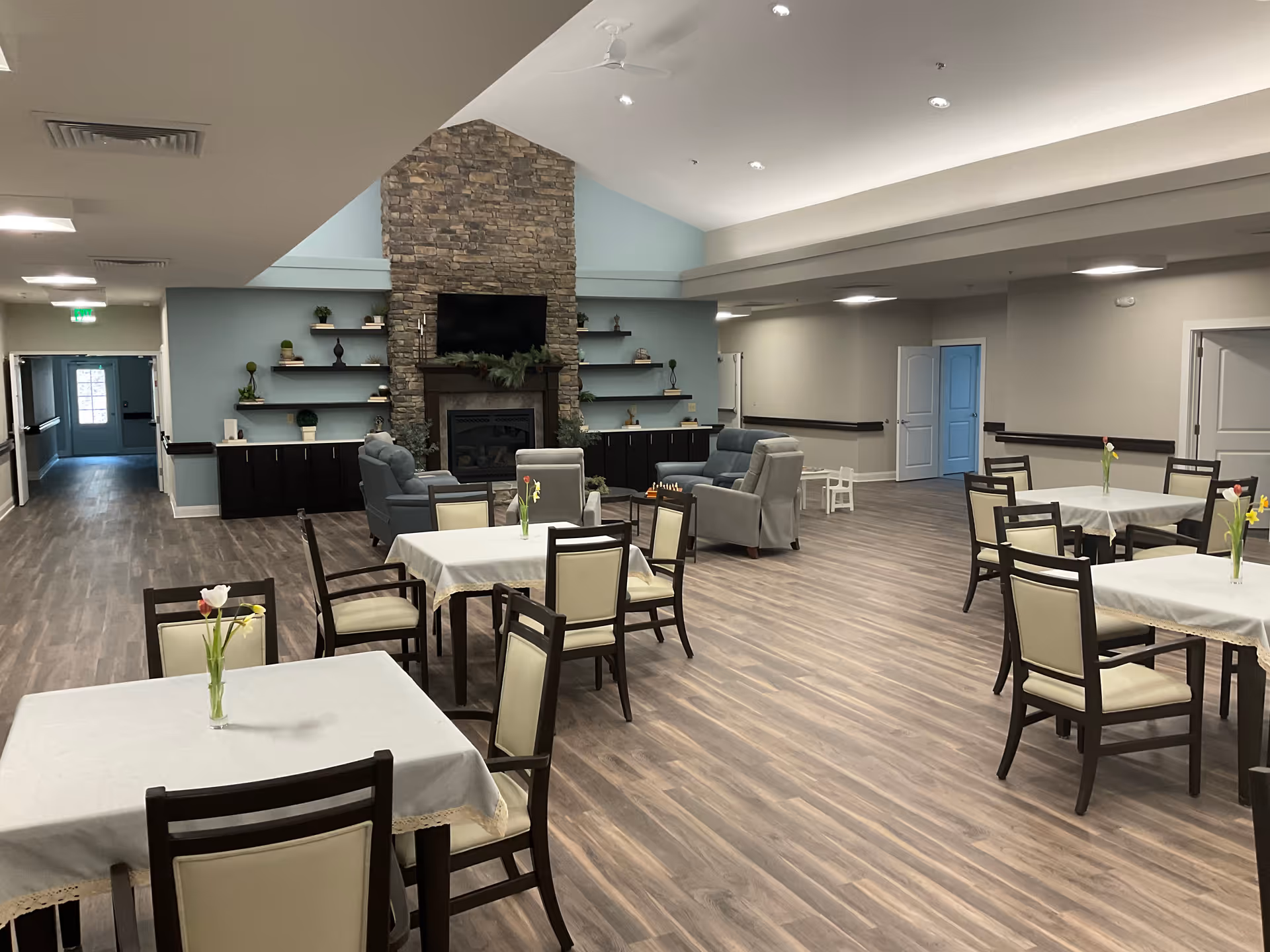 A spacious common area in a senior living facility featuring several tables with chairs covered in white tablecloths and small vases with flowers. In the background, there is a stone fireplace with a mounted TV above it, flanked by shelves with decorative items. Comfortable armchairs are arranged near the fireplace. The room has wood flooring and soft lighting.