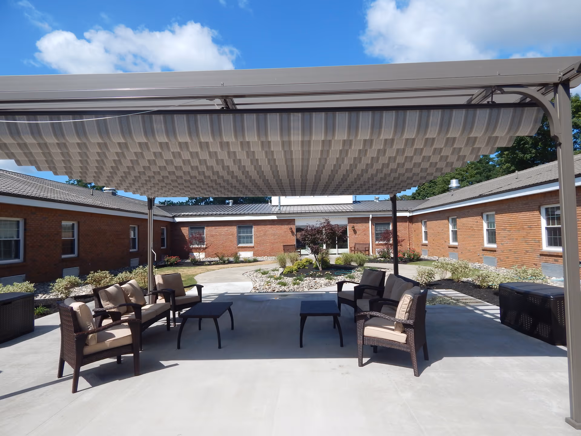 Outdoor seating area at Kingsway Arms Nursing Center with cushioned chairs and small tables under a large retractable awning, surrounded by brick buildings and landscaped garden beds.