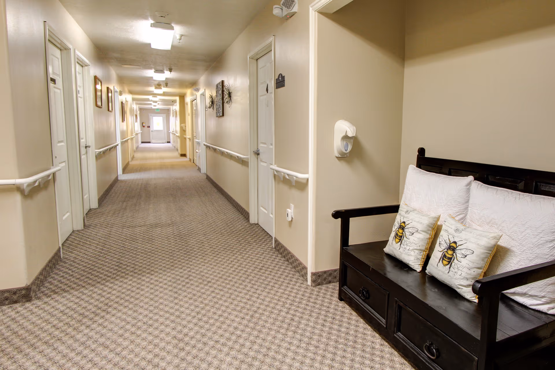 Long carpeted interior hallway with doors on both sides and a bench with bee-themed pillows near the right foreground.