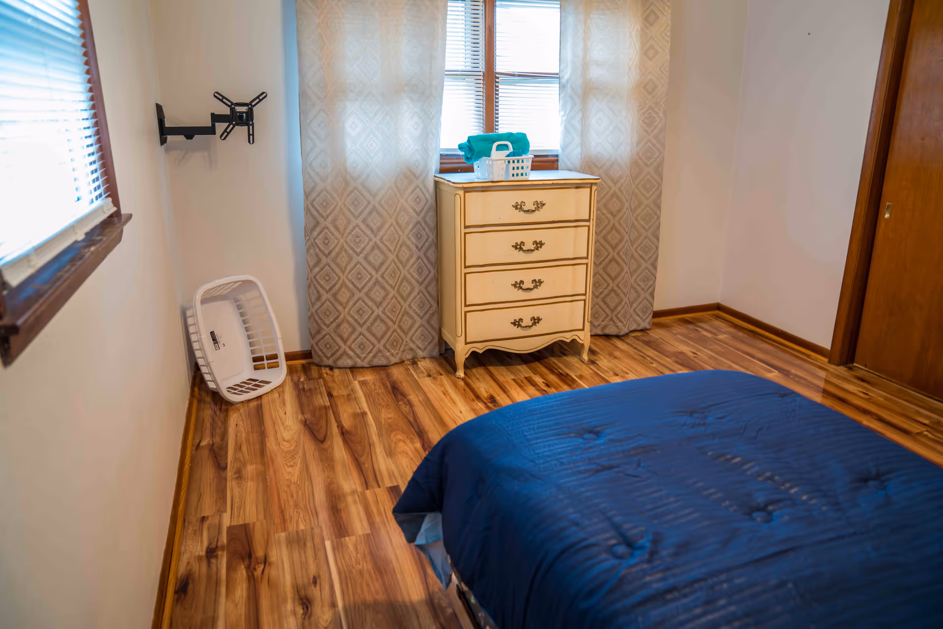 A bedroom with wooden flooring, a bed with a blue comforter in the foreground, a vintage cream-colored dresser with four drawers against the wall, two windows with patterned curtains behind the dresser, a white laundry basket leaning against the wall, and a TV mount bracket on the left wall.