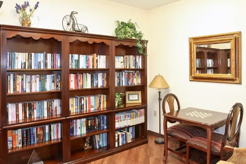 A cozy interior room featuring a large wooden bookshelf filled with books, a small wooden table with a chessboard pattern on top, two wooden chairs with striped cushions, a floor lamp, a gold-framed mirror on the wall, and decorative plants on top of the bookshelf.