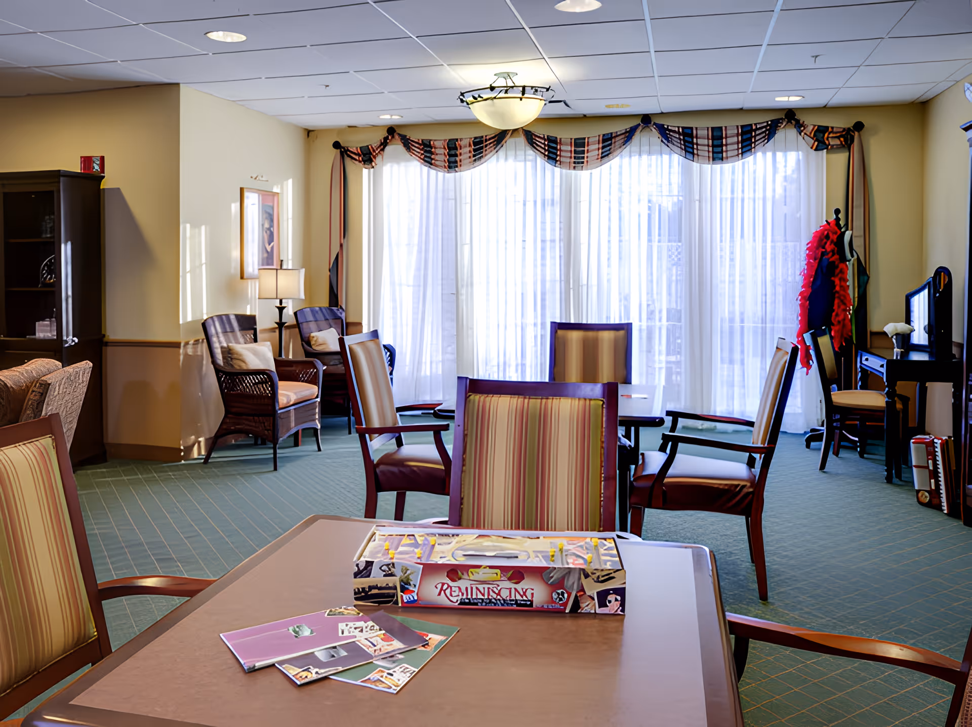 A well-lit common area in a senior living facility with several chairs arranged around tables. A board game box labeled 'Reminiscing' and some booklets are on the table in the foreground. The room has large windows with sheer curtains and decorative valances, a lamp, a coat rack with colorful accessories, and a cabinet with glassware.