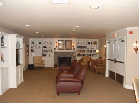A spacious living room area with beige walls and carpet, featuring three brown leather armchairs in the center facing a stone fireplace with built-in bookshelves on either side. Additional seating includes two armchairs and a small table with a lamp near the fireplace. The room is softly lit with ceiling lights and wall sconces, and there are double doors on the right side of the image.