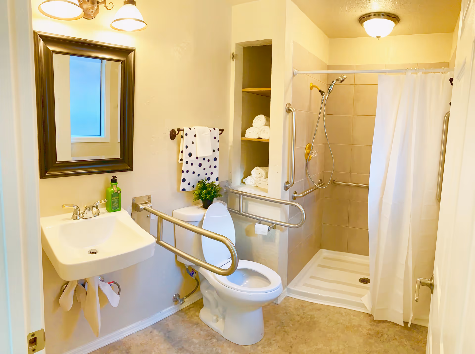 A well-lit bathroom featuring a white toilet with safety grab bars on both sides, a wall-mounted sink with a mirror above it, a towel rack holding a white towel with black polka dots, and an open shower area with a handheld showerhead and grab bars. There are shelves built into the wall stocked with rolled white towels, and a small green plant is placed on the toilet tank.