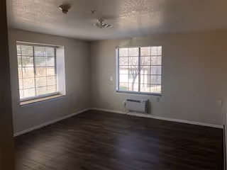 Empty room with two windows letting in natural light, beige walls, dark wood flooring, a ceiling light fixture, and a wall-mounted air conditioning unit below one window.