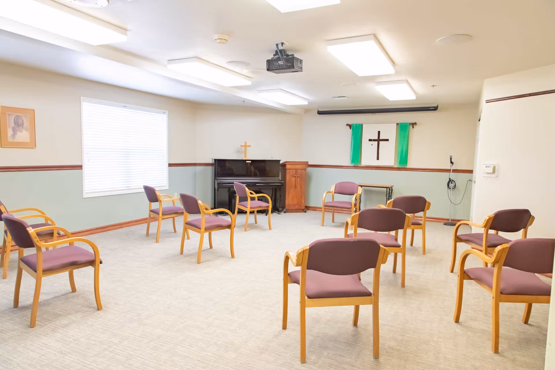A small chapel or meeting room with wooden chairs arranged in rows facing a piano and a wooden podium with a cross. The room has light-colored walls with a green lower half and a picture of Jesus on one wall. There is a cross with green cloth hanging on the wall behind the podium and a microphone stand to the right.
