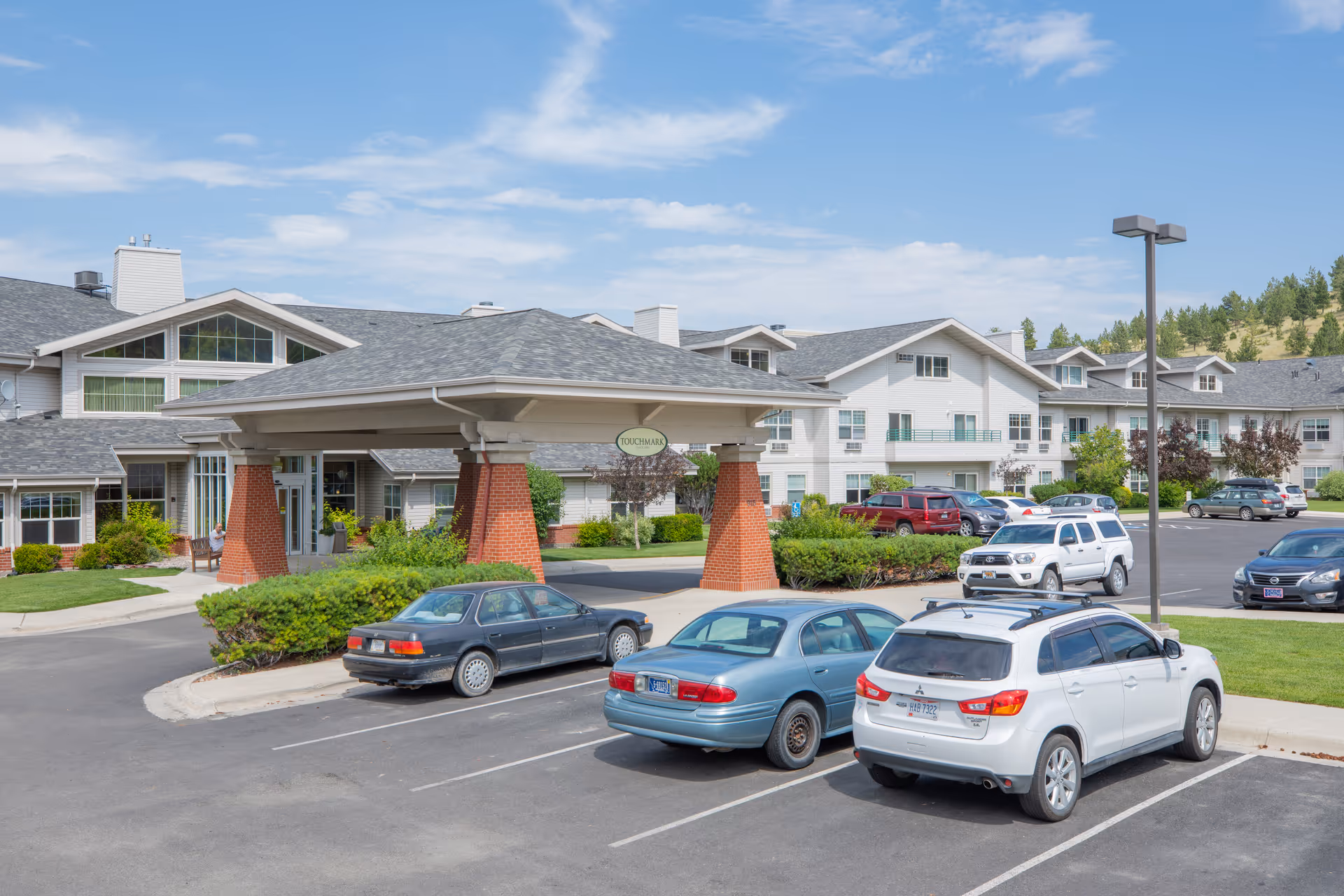 Exterior view of Touchmark on Saddle Drive senior living facility showing the main entrance with a covered drop-off area supported by brick pillars. Several cars are parked in the parking lot in front of the building. The building is white with multiple windows and a gray roof, surrounded by green landscaping and trees under a partly cloudy sky.