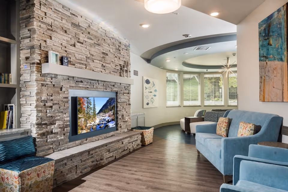 Modern communal lounge with a stone fireplace and mounted TV, blue upholstered seating, and large bay windows.
