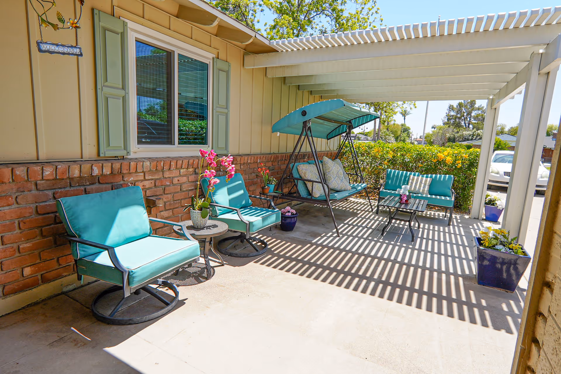 Outdoor patio area with turquoise cushioned chairs, a swing bench with pillows, small tables with potted flowers, and a pergola casting striped shadows on the concrete floor. The patio is adjacent to a brick and siding building with a window and green shutters, surrounded by greenery and parked cars visible in the background.