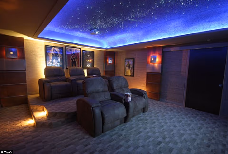 A cozy home theater room with two rows of plush reclining chairs facing a screen. The ceiling is decorated with blue lighting and star-like patterns, creating a night sky effect. The walls have framed movie posters and soft wall sconces emitting warm light. There is a small step with lights leading to the back row of seats.