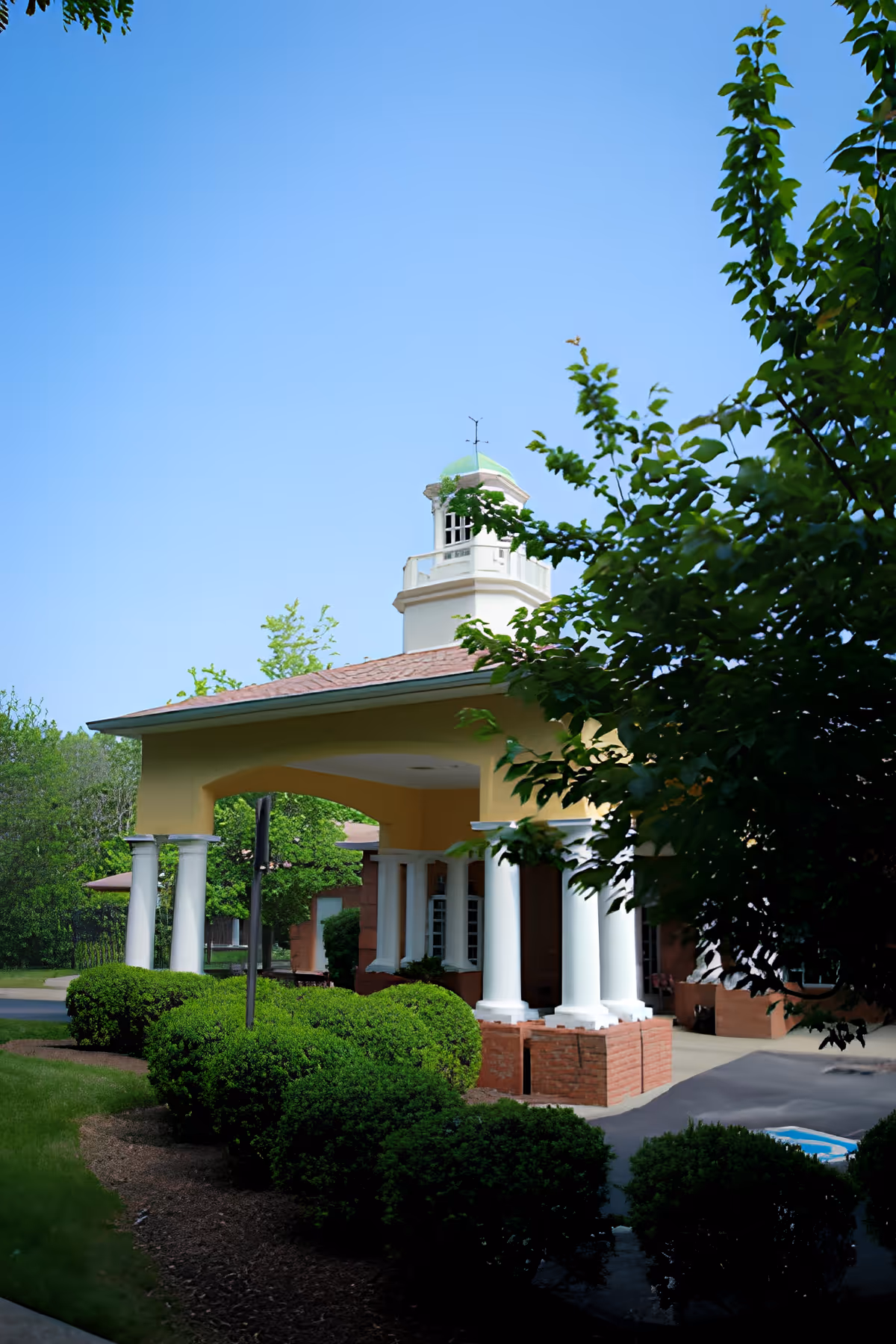 Exterior view of a building entrance with white columns and a yellow overhang, surrounded by green bushes and trees under a clear blue sky.