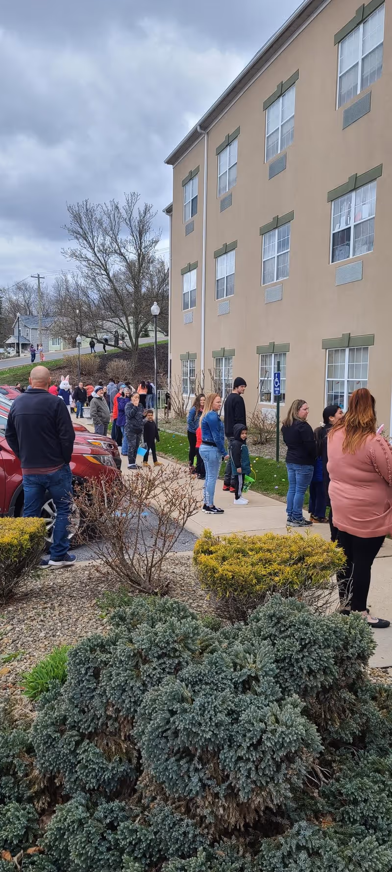 People lined up on the sidewalk outside a multi-story beige nursing facility under a cloudy sky.