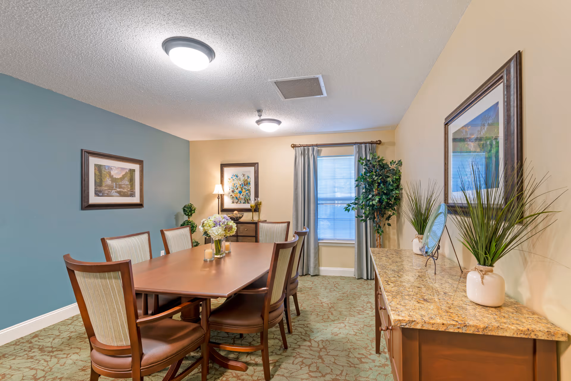 A dining room with a wooden table surrounded by six chairs. The room has a green carpet with a floral pattern, a blue accent wall with a framed picture, and beige walls with another framed artwork. There is a window with gray curtains, a sideboard with a granite top decorated with plants and a decorative plate, and a small chest of drawers with a lamp and decorative items. The ceiling has two round light fixtures.