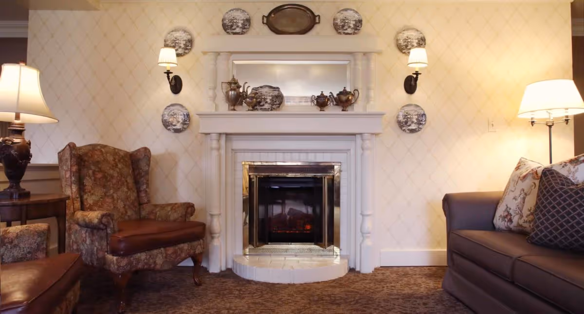 A cozy living room area featuring a white fireplace with decorative plates and silver tea sets on the mantel. To the left is a floral upholstered armchair with a brown leather seat cushion next to a wooden side table with a lamp. To the right is a dark gray sofa with patterned throw pillows and a floor lamp beside it. The walls have a subtle diamond pattern wallpaper.