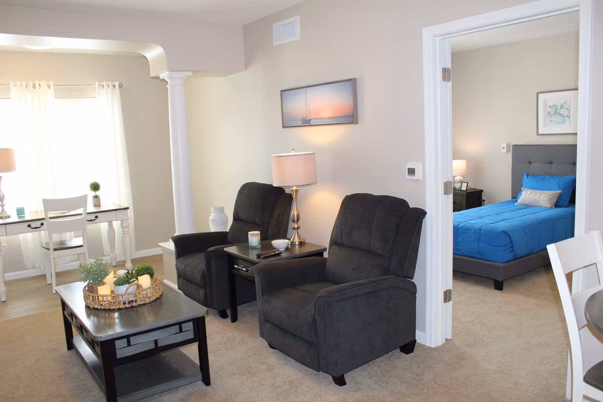 A bright living room with two gray recliners, a coffee table, side table with lamp, and a view into a bedroom with a blue bed.