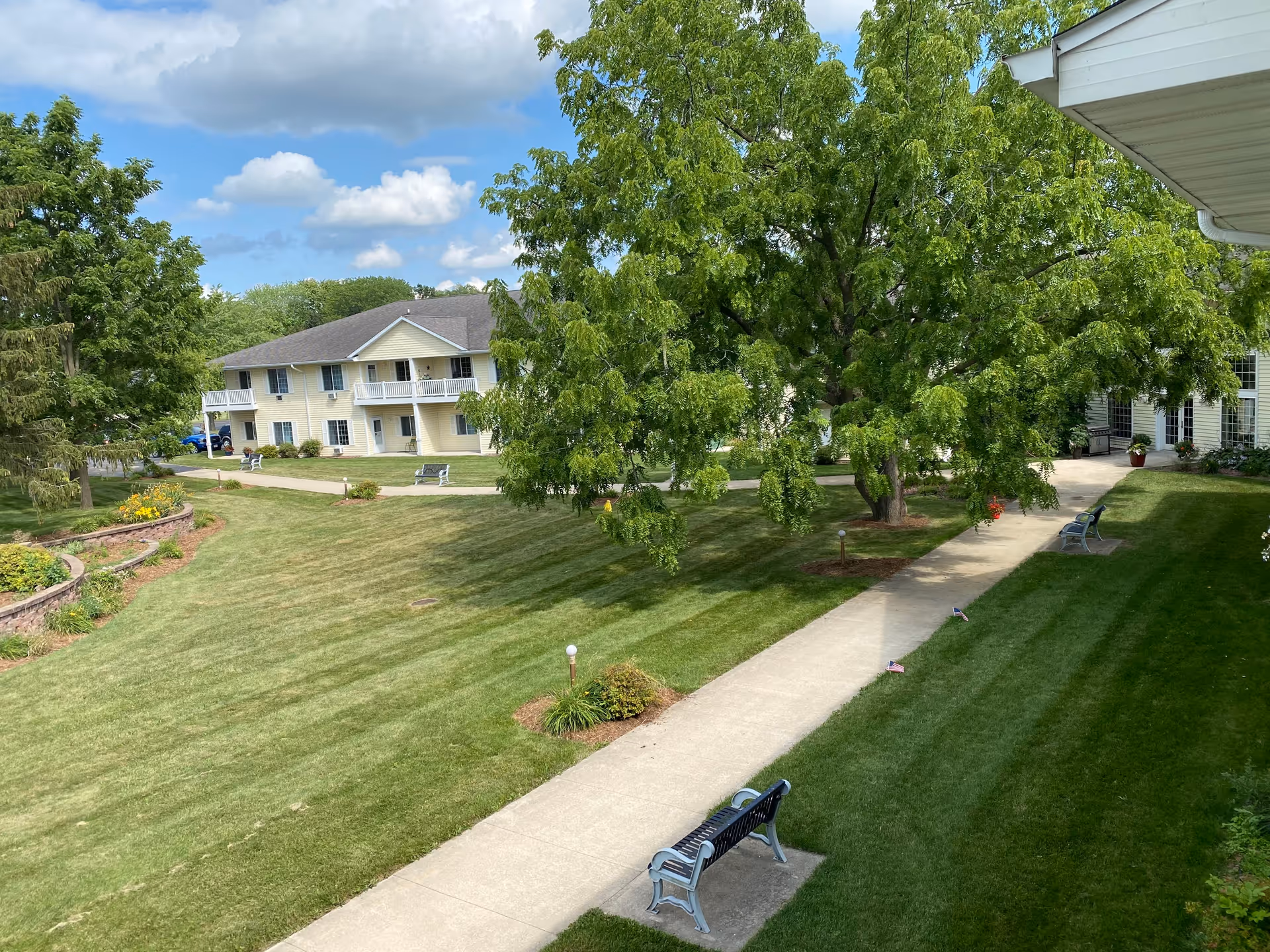 A well-maintained outdoor area of Charleston House Assisted Living featuring a paved walkway, green lawns, several benches, large leafy trees, and a two-story building with balconies in the background under a partly cloudy sky.