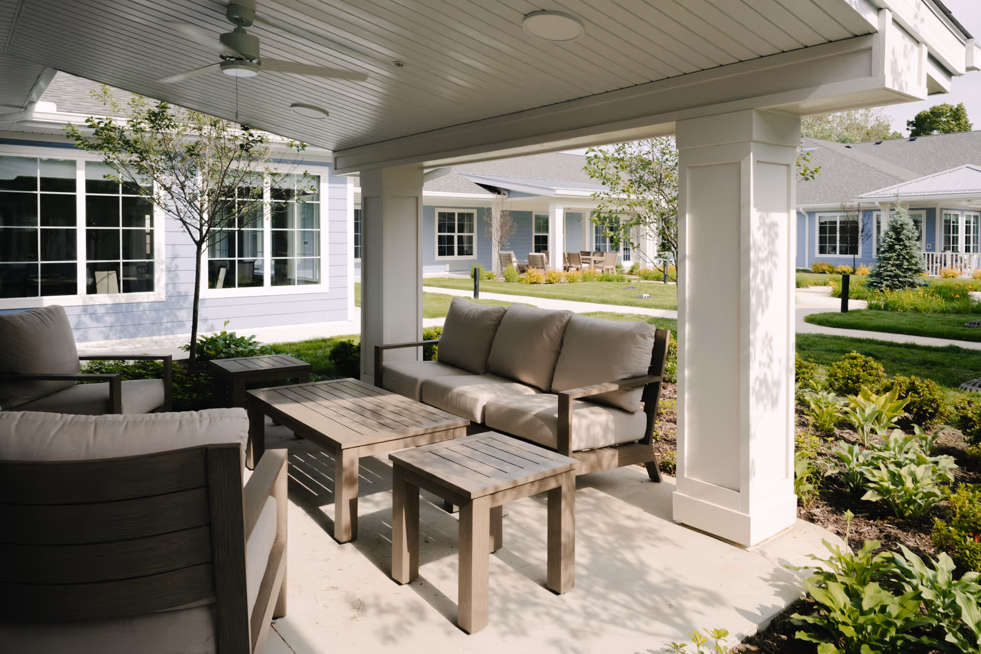 Outdoor covered seating area with cushioned wooden chairs and tables, surrounded by landscaped garden beds and pathways, with light blue residential buildings in the background.