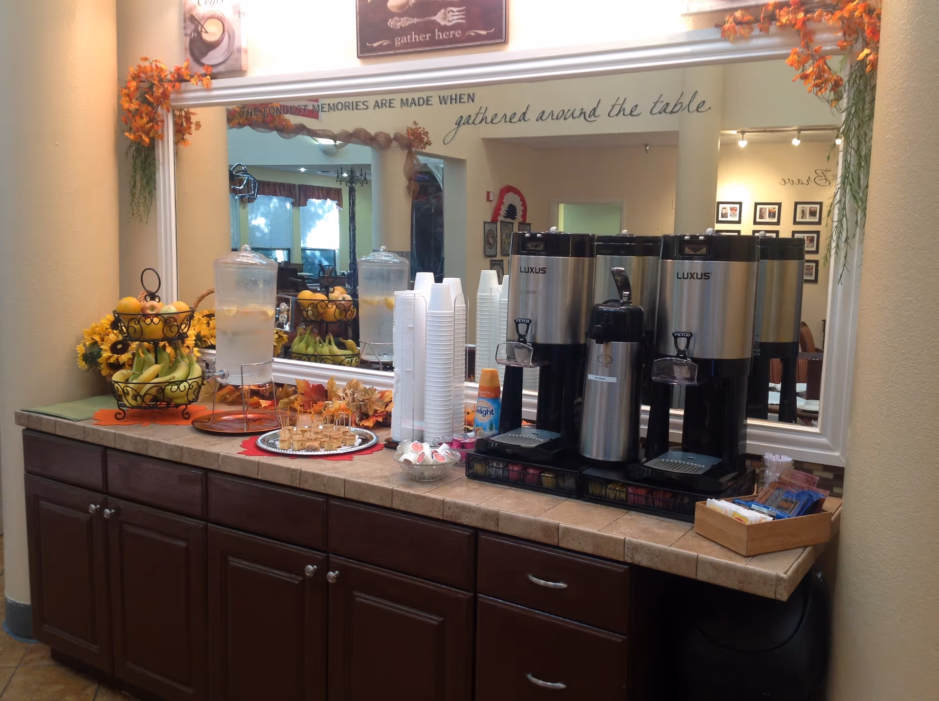 A beverage and snack station in a retirement community with two large water dispensers, stacks of white disposable cups, a tiered fruit basket with bananas and apples, a plate of small sandwiches, and a container of coffee creamers. The station is set on a tiled countertop with dark wooden cabinets below. Above the station is a large mirror decorated with autumn leaves and a sign that reads 'The fondest memories are made when gathered around the table.'
