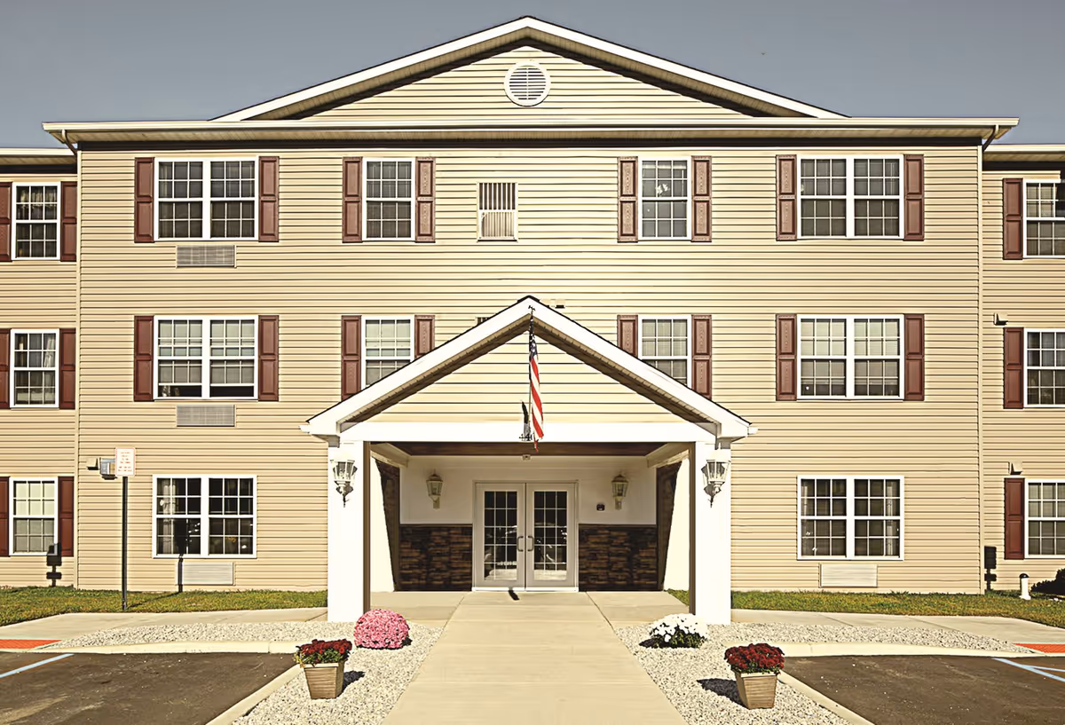 Front exterior view of a three-story senior apartment building with beige siding and maroon shutters. The entrance has a covered porch with white pillars and an American flag hanging above the double glass doors. There are flower pots with red, pink, and white flowers along the walkway leading to the entrance.