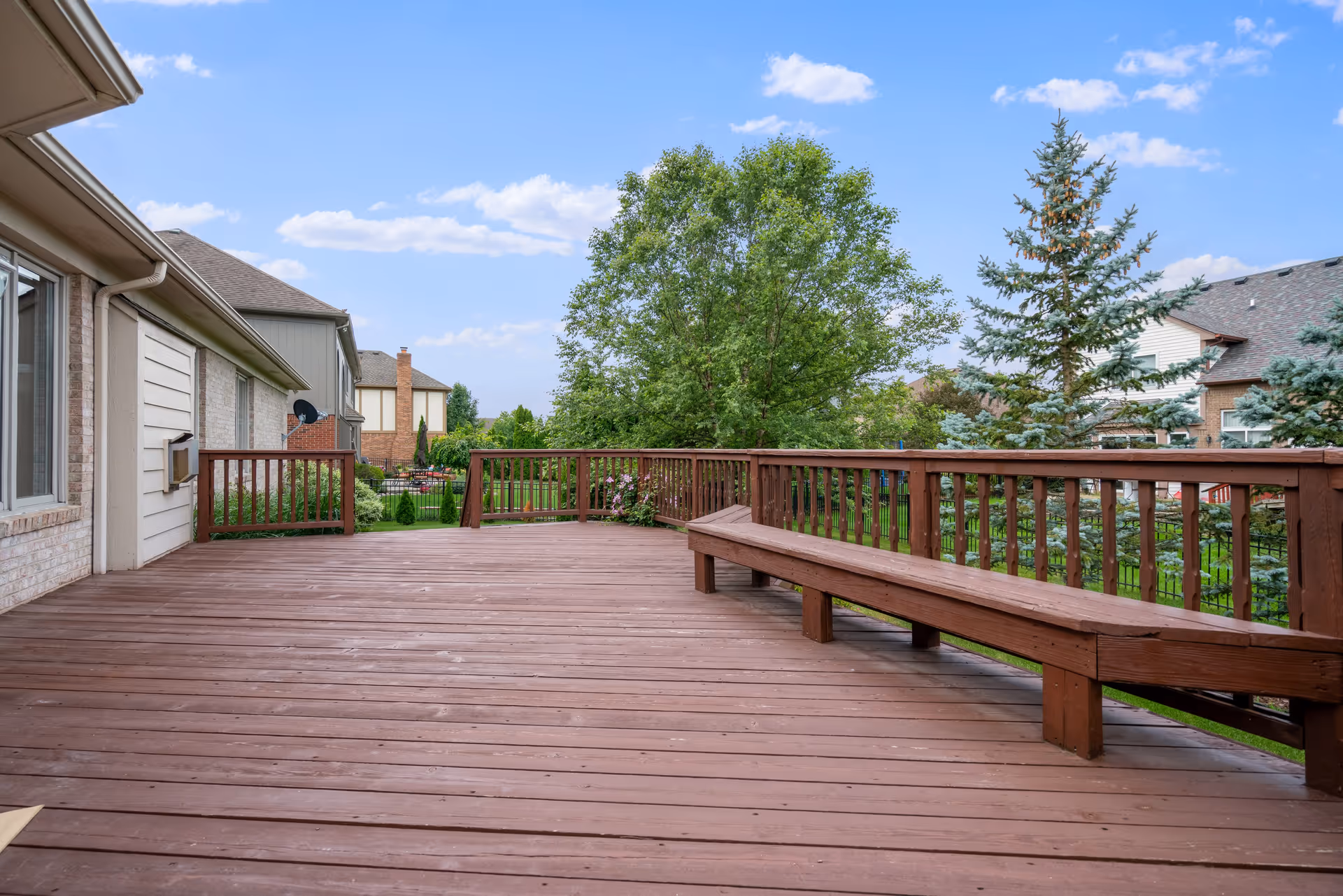A spacious wooden outdoor deck attached to a house, featuring built-in wooden bench seating along the railing. The deck overlooks a green backyard with trees and neighboring houses under a partly cloudy blue sky.