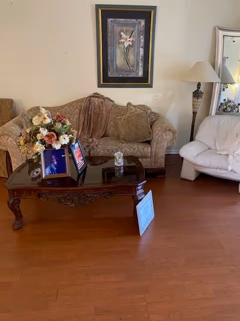 A cozy living room area with a patterned beige sofa, a white cushioned chair, a wooden coffee table with floral arrangement and framed photos, a floor lamp, a large mirror, and a framed floral painting on the wall.