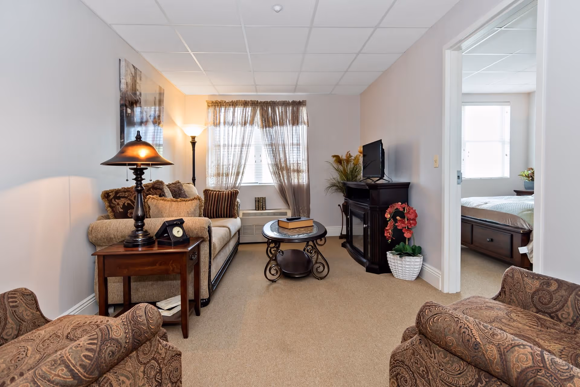 A cozy living room with a beige sofa adorned with multiple cushions, a wooden side table with a lamp and clock, a round coffee table, and two patterned armchairs. A window with sheer curtains lets in natural light. A flat-screen TV sits on a dark wooden cabinet next to a potted plant with red flowers. An open doorway reveals a bedroom with a bed and a window.