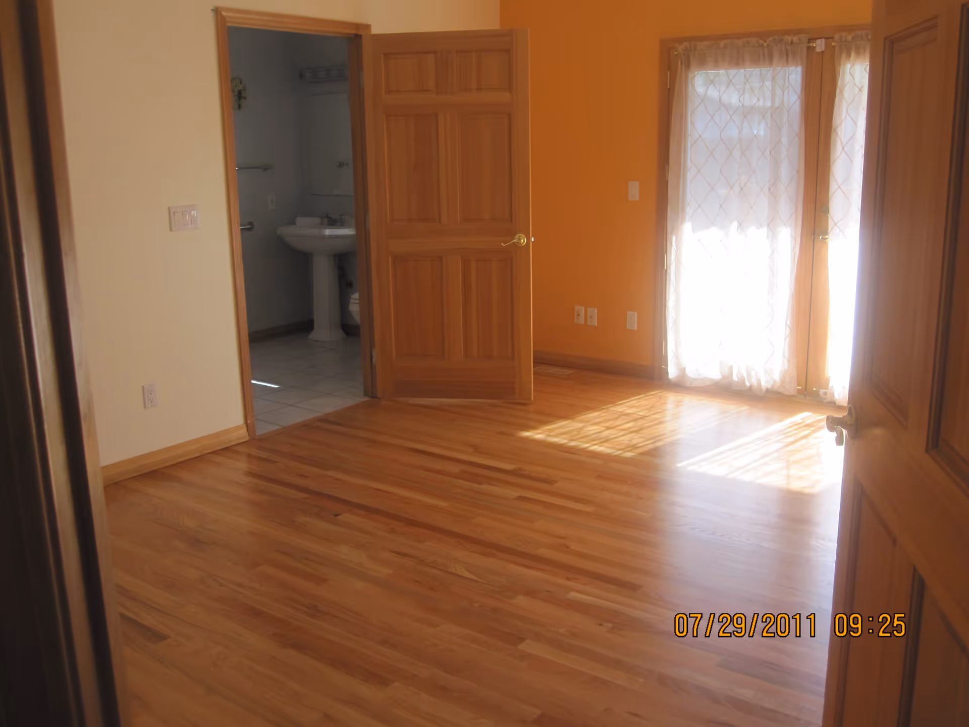 Empty room with polished wooden floor, light yellow and orange walls, an open wooden door leading to a bathroom with a pedestal sink and toilet, and glass doors with sheer curtains allowing sunlight to enter.