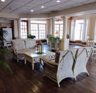 A bright and spacious living room area with wicker furniture including a sofa and chairs with floral cushions, a glass-top coffee table with a lantern centerpiece, large windows allowing natural light, wooden flooring, and decorative plants.