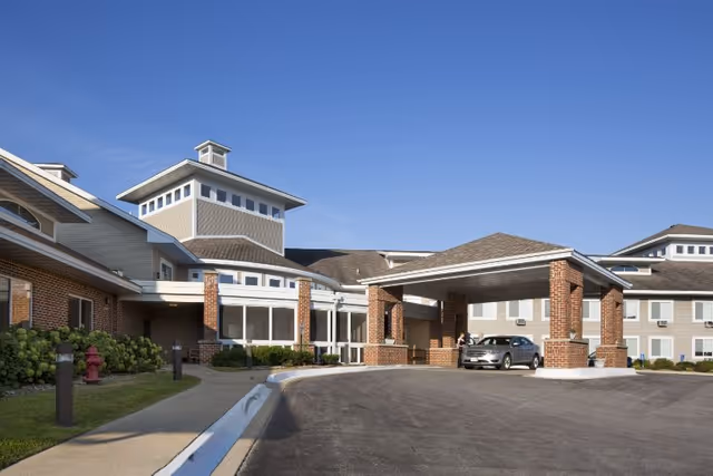 Front entrance of a brick-faced assisted living facility with a covered drop-off porte-cochere, a parked car, and clear blue sky.
