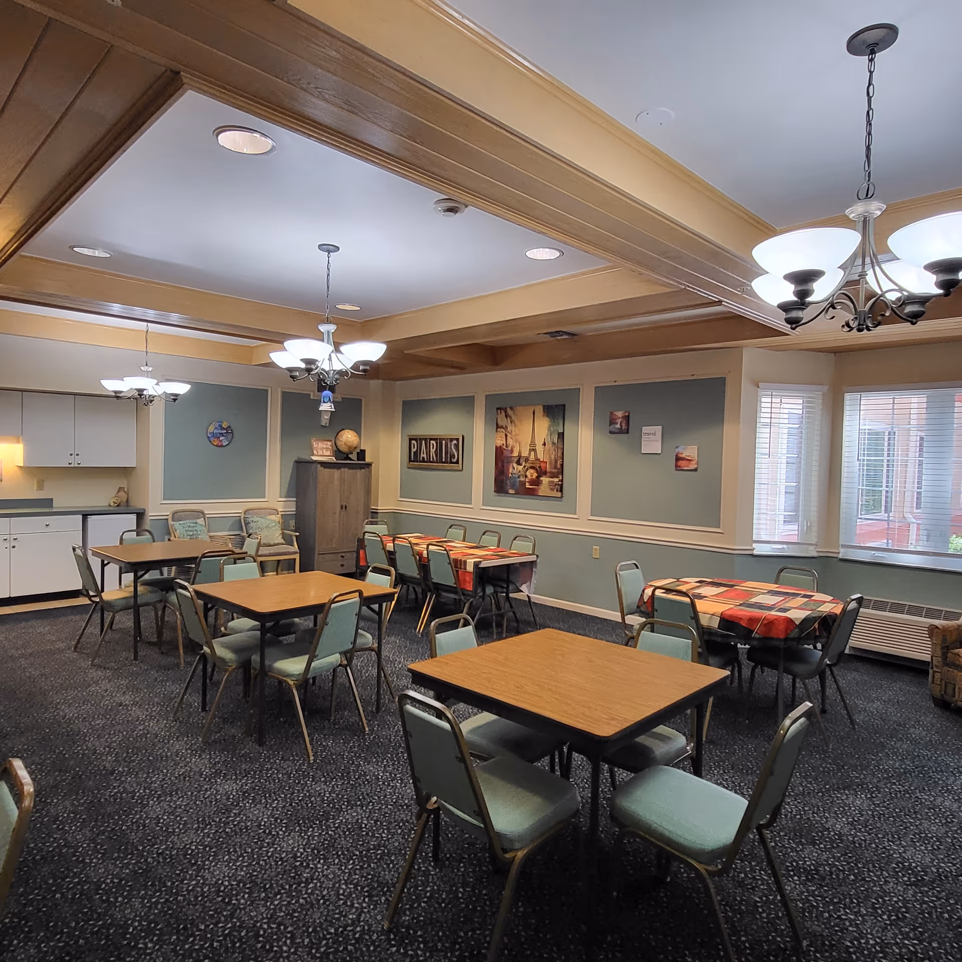 A dining room with multiple square wooden tables and green cushioned chairs arranged around them. Two tables have red and beige checkered tablecloths. The room has blue walls with white trim, decorated with framed pictures including one of the Eiffel Tower and a sign that says 'PARIS'. There are two chandeliers hanging from the ceiling, and windows with white blinds letting in natural light. A cabinet and some additional chairs are placed against the walls.