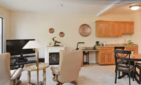 A cozy living area in Saratoga Retirement Community featuring two beige armchairs around a small glass-top table with a lamp, a flat-screen TV on a stand, a white electric fireplace, and a wooden dining table with black chairs. The room has light-colored walls, carpeted floor, and wooden cabinets with a countertop along one wall.