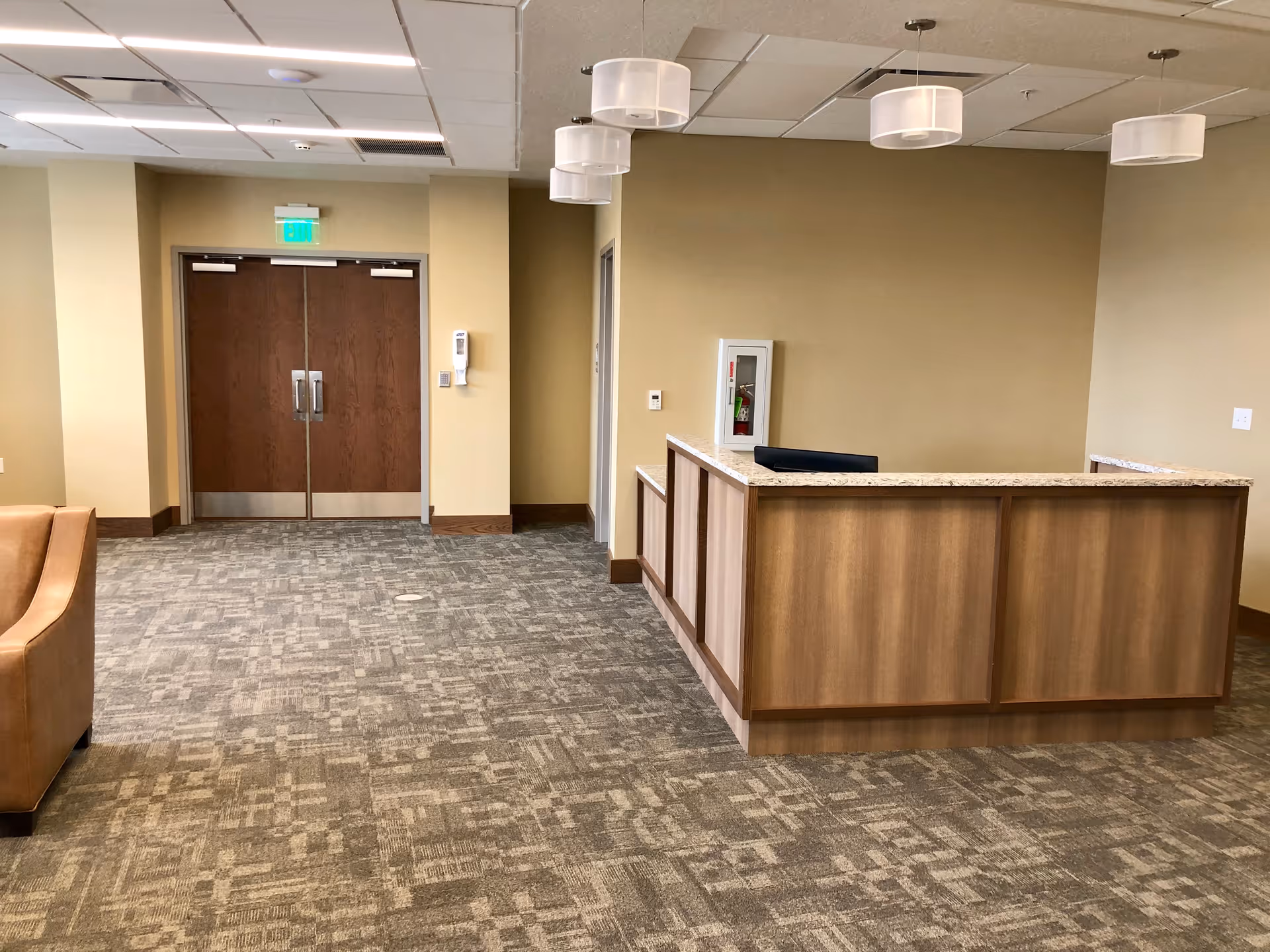 Interior view of a reception area in an assisted living facility featuring a wooden reception desk with a granite countertop, beige walls, patterned carpet flooring, a brown leather chair partially visible on the left, and double wooden doors with metal handles in the background.