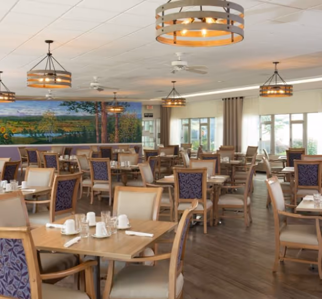 A spacious dining room with multiple wooden tables and chairs arranged neatly. Each table is set with white cups, glasses, and napkins. The chairs have beige seats and backs with a purple patterned fabric on the backrest. The room features large windows with beige curtains allowing natural light to enter. The ceiling has several modern circular light fixtures and ceiling fans. A large mural depicting a scenic landscape with trees and a field is visible on one wall.