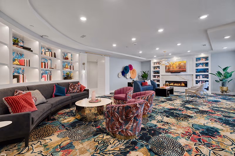 Bright, colorful communal lounge with a curved sofa, patterned chairs, built-in bookshelves, a fireplace, and a vibrant patterned carpet.