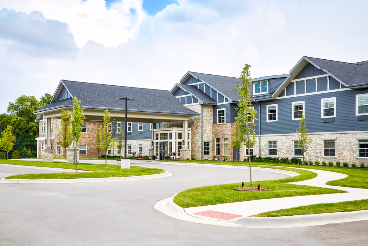 Exterior view of Provision Living at East Lansing, showing a modern two-story building with a covered entrance, stone and blue siding, multiple windows, young trees, and a curved driveway with well-maintained green lawns.