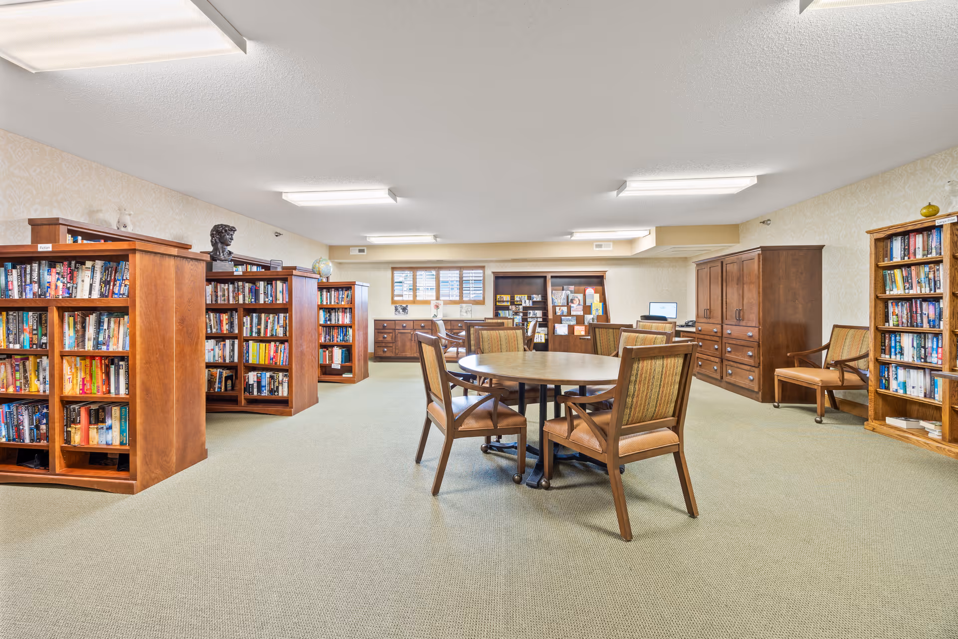 A spacious library room with multiple wooden bookshelves filled with books, a round table surrounded by four wooden chairs with cushioned seats, and additional wooden furniture including cabinets and chairs along the walls. The room is well-lit with ceiling lights and has a beige carpet and wallpaper.