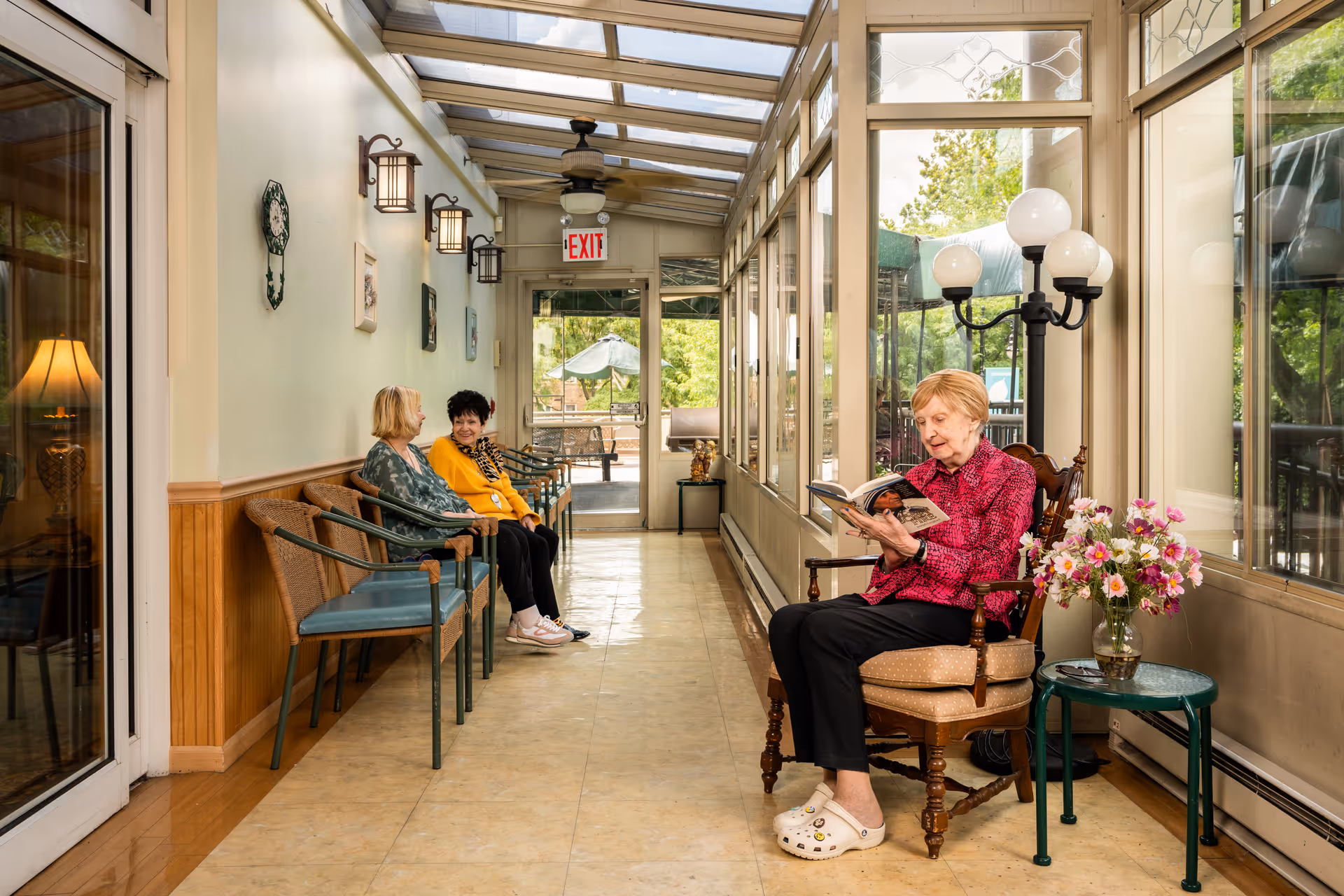 A sunlit hallway with large windows on one side and a row of chairs along the opposite wall. Two elderly women are seated together chatting, while another elderly woman sits alone reading a book in a cushioned wooden chair next to a small table with a vase of flowers. The hallway has a tiled floor, wall-mounted lamps, and an exit door at the end leading outside.
