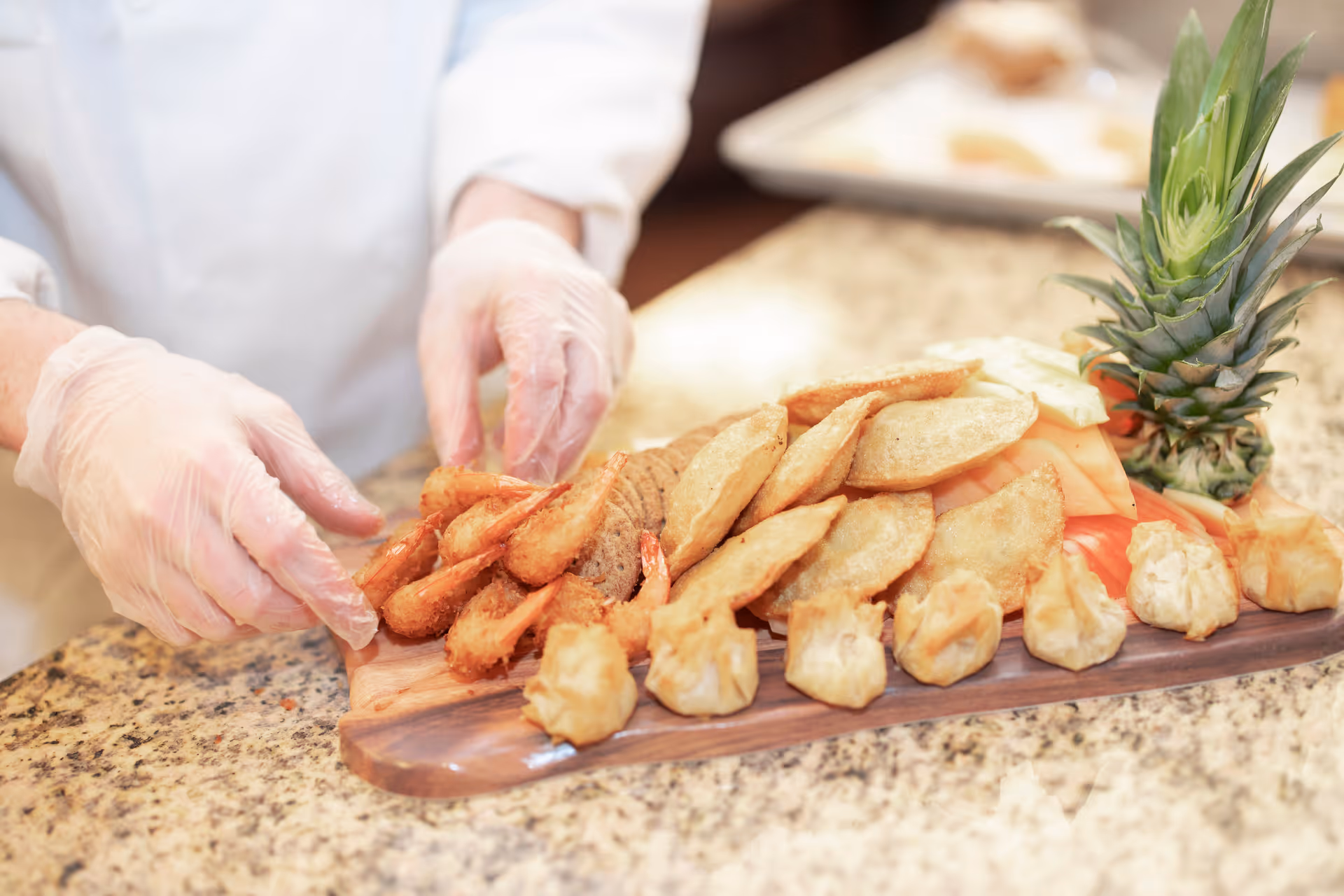 A person wearing gloves arranges a wooden platter with various fried appetizers including shrimp, potato wedges, and dumplings, garnished with a pineapple top and slices of melon on a granite countertop.