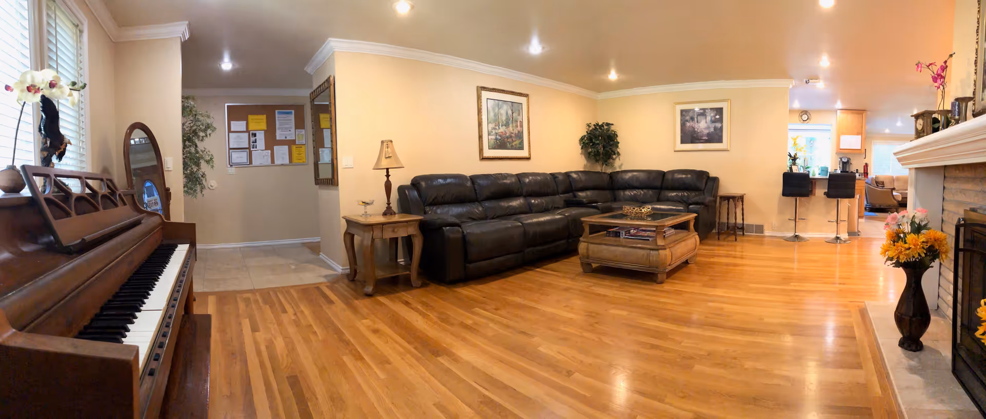 A cozy living room with wooden flooring, a large black leather sectional sofa, a wooden coffee table, and a side table with a lamp. There is a piano on the left side near a window with white blinds and a decorative orchid on top. The room has beige walls with framed artwork and a fireplace on the right with a vase of flowers. In the background, there is a kitchen area with two black bar stools and some plants.