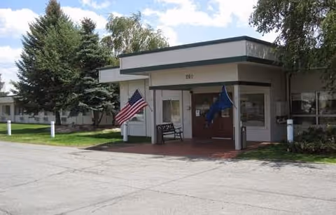 Front entrance of a single-story building with two flags, one American and one blue, displayed on either side of the doorway. There is a bench near the entrance and trees and grass surrounding the building under a partly cloudy sky.