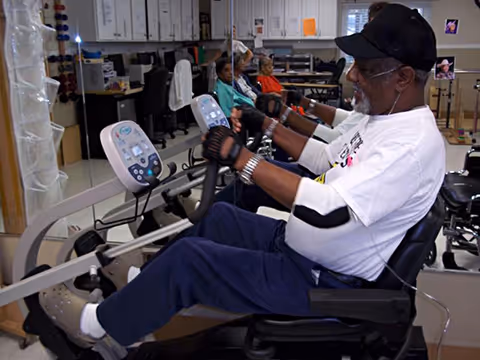 An elderly man wearing a white shirt, black cap, and gloves is exercising on a seated rowing machine in a fitness or rehabilitation room. In the background, other elderly individuals are seated and observing or waiting their turn. The room has office chairs, desks, and medical or fitness equipment.