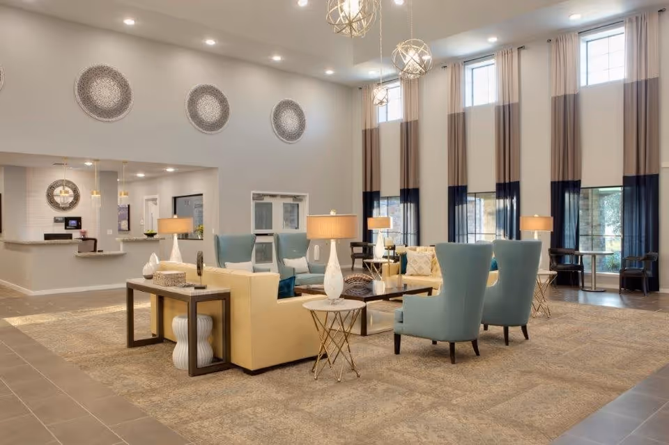 A spacious and well-lit living room area in a senior living facility featuring high ceilings with large windows covered by long curtains. The room is furnished with light blue armchairs, beige sofas, side tables with lamps, and decorative wall plates. The floor is carpeted, and there is a reception desk visible in the background.