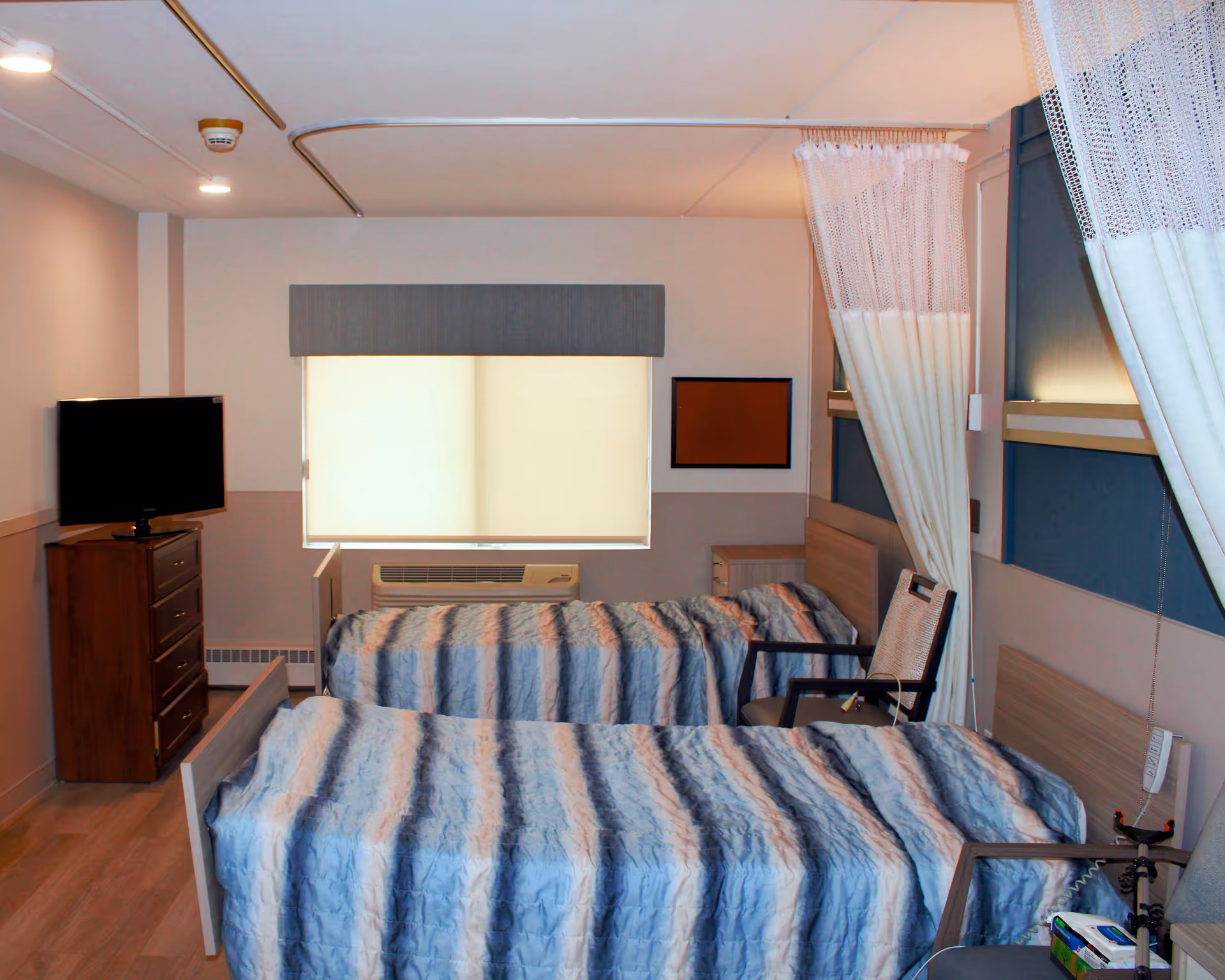 A room with two single beds covered with blue and gray striped blankets, a wooden dresser with a TV on top, a window with a closed blind, a chair between the beds, and privacy curtains partially drawn around the beds.