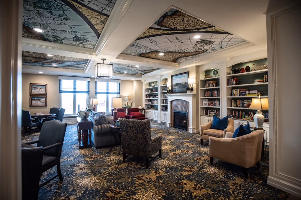 A cozy living room area in a senior living facility featuring patterned carpet and ceiling panels with a vintage map design. The room includes multiple upholstered chairs and sofas arranged around a fireplace with built-in bookshelves on either side. Large windows allow natural light to fill the space, and there are table lamps and framed artwork on the walls.