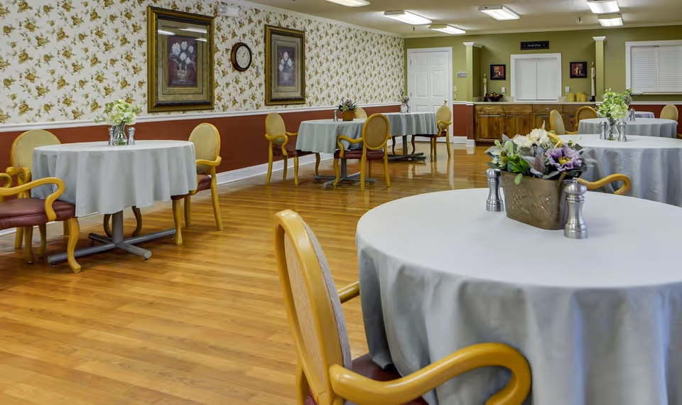A dining room with round tables covered in light gray tablecloths, each table decorated with a small floral centerpiece and salt and pepper shakers. The room has wooden flooring, floral wallpaper with a red lower border, framed artwork, and a clock on the wall. Several wooden chairs with cushioned seats are arranged around the tables. In the background, there is a wooden sideboard with decorative items and a door.