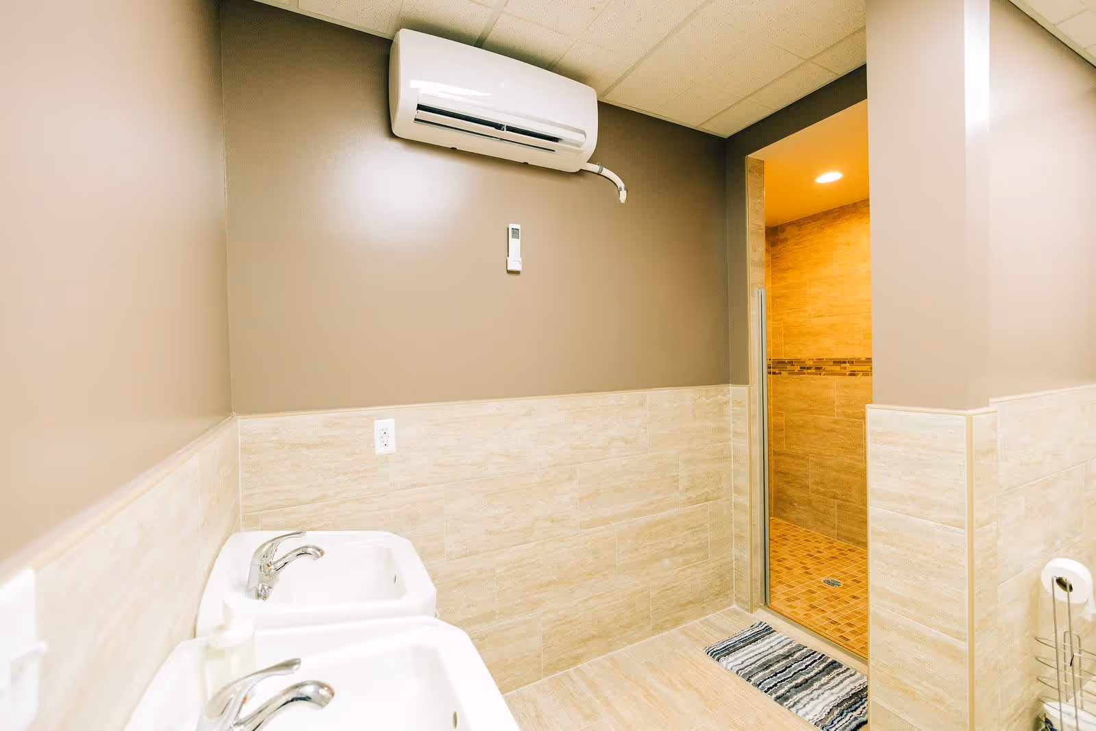 Interior view of a bathroom with two white sinks and chrome faucets on the left, beige tiled walls and floor, a striped rug in front of a walk-in shower with glass door, and a white air conditioning unit mounted on the wall above.