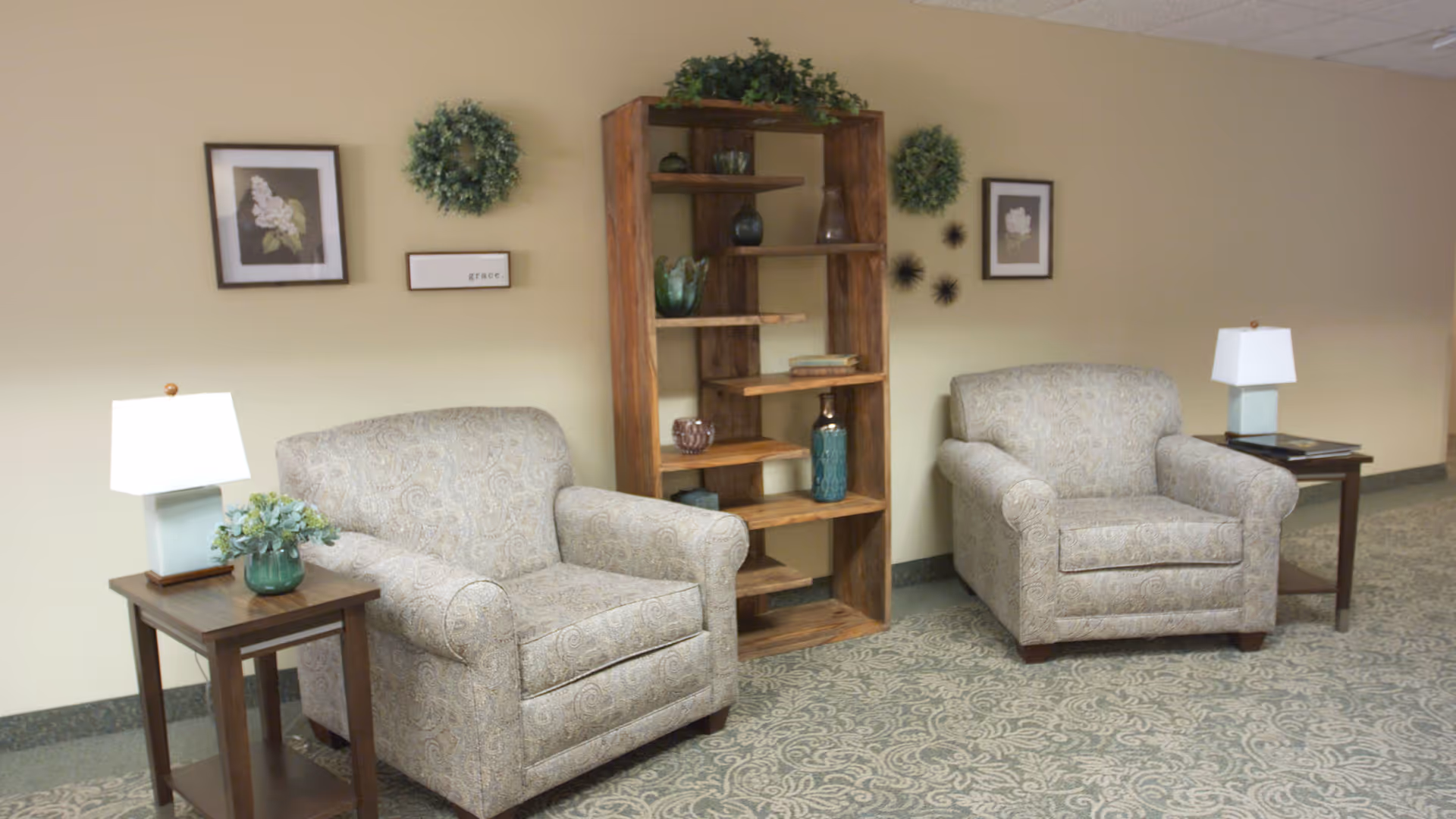 Two patterned armchairs with side tables and lamps beside a wooden bookshelf in a furnished lounge area.